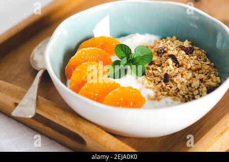 Yogurt bianco con spicchi di tangerina, muesli e foglie di menta in una ciotola bianca e azzurra, con un cucchiaio, su un vassoio di legno Foto Stock