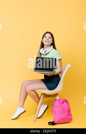 Un adolescente sorridente con computer portatile, cuffie e zaino seduti sulla sedia su sfondo giallo Foto Stock