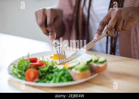 Donna africana irriconoscibile che mangia gustosa colazione in cucina, usando forchetta e coltello Foto Stock