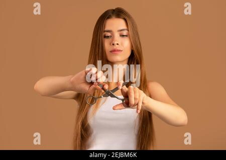 Bella donna millenaria con capelli lunghi perfetti cercando di rompere le forbici su sfondo marrone, fuoco selettivo Foto Stock