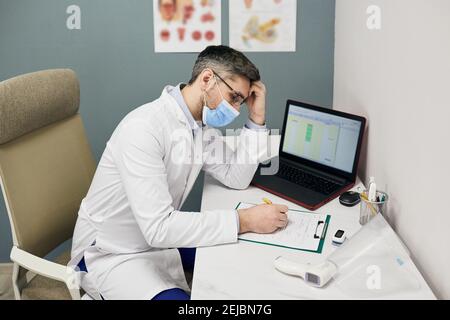 Il medico maschile serio compila il modulo di registrazione medica sul suo posto di lavoro in una clinica medica. Medico indossando camice medico bianco e una maschera protettiva Foto Stock