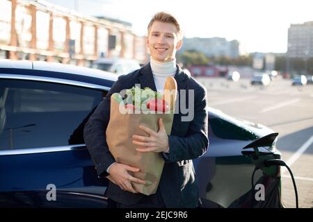 Felice giovane adulto sorridente 30-età uomo che carica la sua auto elettrica, in posa alla macchina fotografica con grande borsa di carta pieno di cibo sano Foto Stock