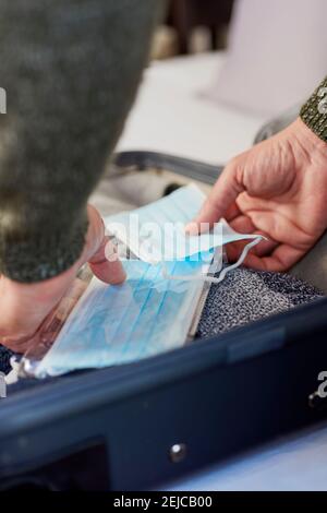 primo piano di un giovane caucasico che sta imballando alcune maschere blu usa e getta nella sua valigia, imballato con diversi vestiti, sul letto Foto Stock