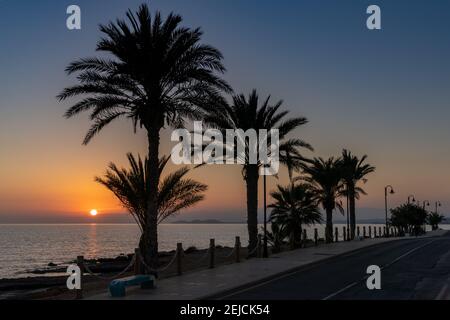 Tramonto colorato sull'oceano con palme in silhouette e un marciapiede sul fronte spiaggia e una strada fronte oceano Foto Stock