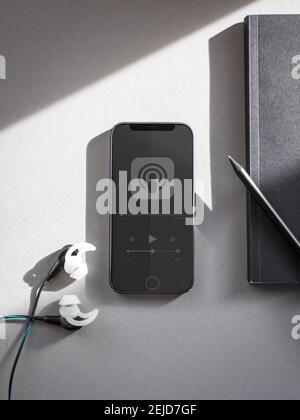 Telefono nero con un podcast in riproduzione e un notebook e auricolari accanto. Foto Stock