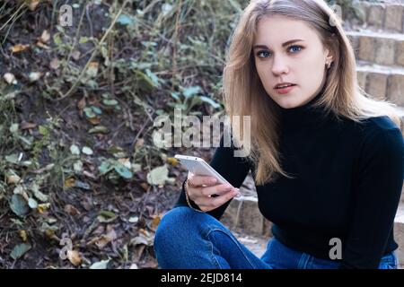Ritratto di una giovane bella donna con gli occhi blu un dolcevita nero che si affaccia sulla fotocamera passi all'esterno tenendo un pho mobile Foto Stock