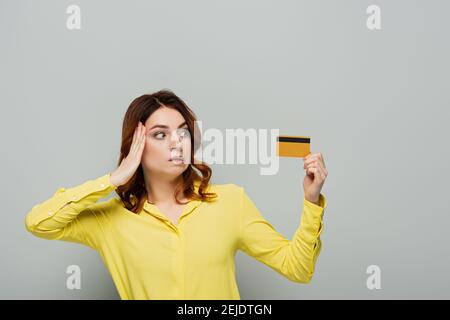 donna bemused che tocca la testa mentre tiene la carta di credito sul grigio Foto Stock