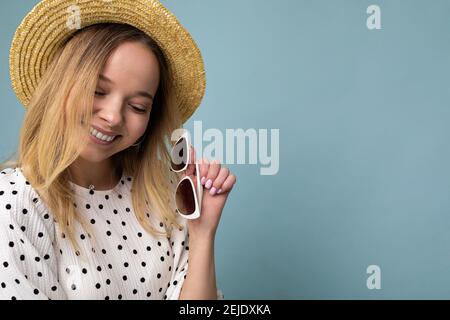 Primo piano di una giovane donna bionda sorridente abbastanza positiva che indossa abito estivo cappello di paglia ed eleganti occhiali da sole isolati su blu parete di fondo Foto Stock