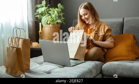 Felice giovane donna disimballare online ordini di merci o cibo. Acquisto online, consegna ordini. Adolescente ragazza rilassarsi sul divano considerando gli acquisti con Foto Stock