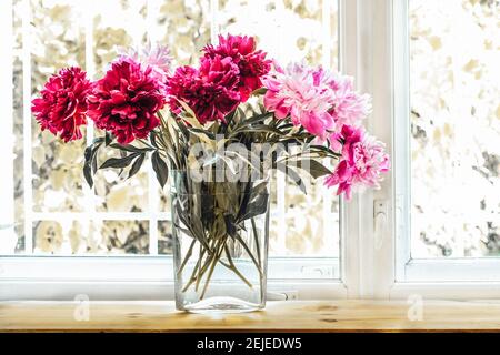 Bouquet di belle fresche e brillanti peonie rosa in vaso trasparente di vetro sullo sfondo della finestra. Immagine con tonalità retrospettiva. Tempo di fioritura. Vacanza vintage co Foto Stock