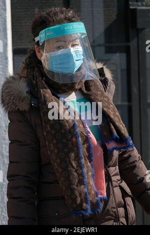 Una donna asiatica cauto americana, probabilmente cinese indossa maschere di traino e uno scudo facciale. In un parco a Flushing, Queens, New York City. Foto Stock