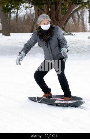 Una donna americana cinese attiva nel suo 50s slitta giù una collina nevosa in un inverno freddo. In un parco a Flushing, Queens, New York City. Foto Stock