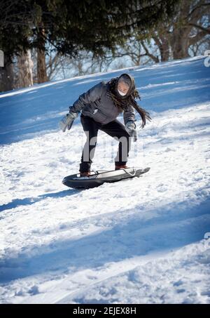 Una donna americana cinese attiva nel suo 50s slitta giù una collina nevosa in un inverno freddo. In un parco a Flushing, Queens, New York City. Foto Stock