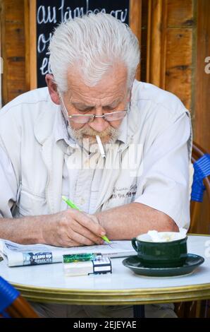 Uomo che ha un cappuccino mattina e fare un puzzle crossword mentre si fuma. Al Cafe Gaby a Lourmarin, Francia Foto Stock