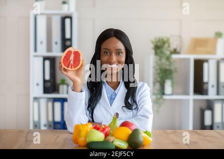 Positivo esperto di nutrizione nera che tiene il pompelmo alla scrivania in medicina ufficio Foto Stock