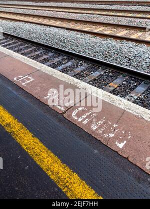 Striscia di avvertimento gialla e mente sbiadita la nota di gradino sulla piattaforma della stazione ferroviaria del Regno Unito con binari ferroviari in background. Foto Stock