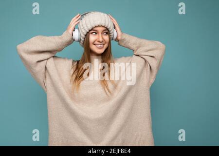Bella felice sorridente giovane donna bionda con maglione invernale beige e cappello isolato su sfondo blu indossando bluetooth wireless bianco Foto Stock