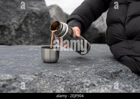 Immagine ritagliata di una donna seduta su rocce che tengono thermos, versa una bevanda calda in una tazza e facendo una pausa mentre camminando in natura. Foto Stock