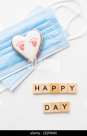 Maschera medica monouso e due cuori. Cuore con footprint del bambino. Parto nella pandemia del coronavirus. Lettere di buon giorno Foto Stock