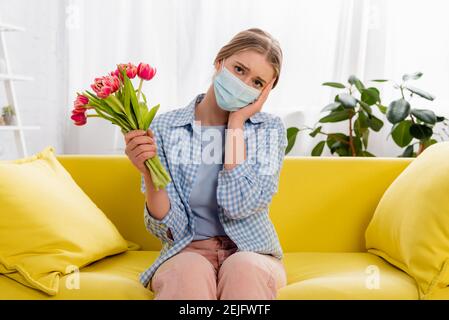 Donna triste in maschera medica che tiene tulipani durante l'allergia Foto Stock