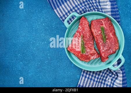 Bistecca cruda su un tagliere con rosmarino e spezie, sfondo blu, vista dall'alto. Carne fresca alla griglia. Bistecca di manzo alla griglia. Foto Stock