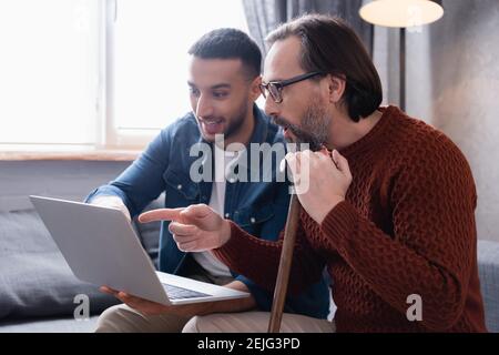 uomo stupito con bastone che punta verso il laptop vicino ai giovani figlio ispanico Foto Stock
