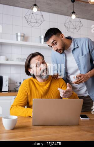 uomo stupito che indicò il computer portatile vicino al figlio ispanico in cucina Foto Stock
