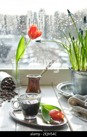 Caffè orientale in tradizionale caffettiera turca di rame con fiori sul davanzale della finestra. Davanzale in legno con tulipani arancioni e vaso di fiori di giacinto Foto Stock