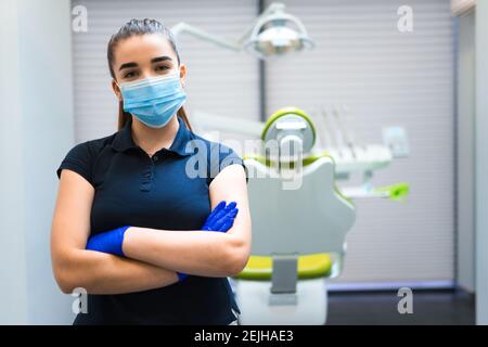 Giovane donna dentista medico in maschera si trova in ufficio con le braccia incrociate Foto Stock