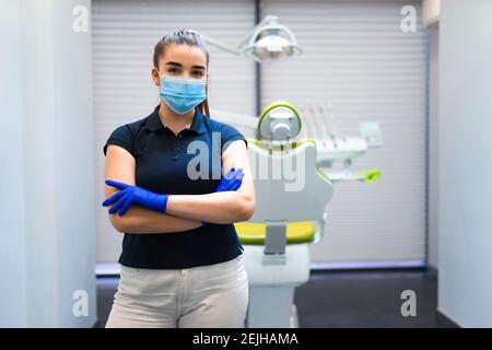 Giovane donna dentista medico in maschera si trova in ufficio con le braccia incrociate Foto Stock