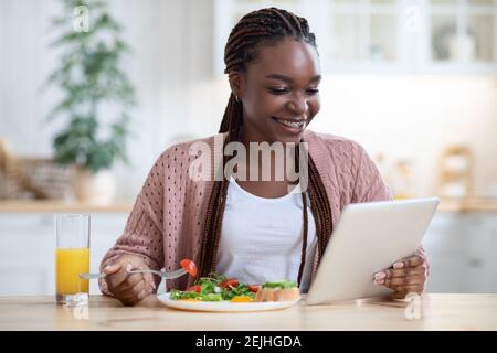 Allegra donna nera con Tablet digitale che legge notizie durante la colazione In cucina Foto Stock