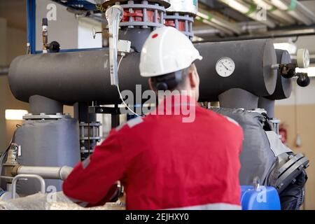 Tema del settore energetico: Un tecnico vestito con tute rosse e un casco bianco che controlla i parametri di riscaldamento. Tecnologia di distribuzione dell'acqua di riscaldamento. Foto Stock