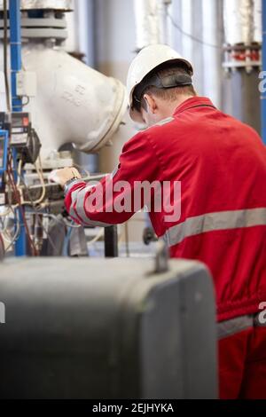 Tema del settore energetico: Un tecnico vestito con tute rosse e un casco bianco che controlla i parametri di riscaldamento. Tecnologia di distribuzione dell'acqua di riscaldamento. Foto Stock