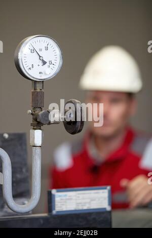 Termometro sul tubo di alimentazione dell'acqua calda, impianto di riscaldamento. Tecnico che indossa una giacca rossa con casco bianco che controlla i parametri. Foto Stock