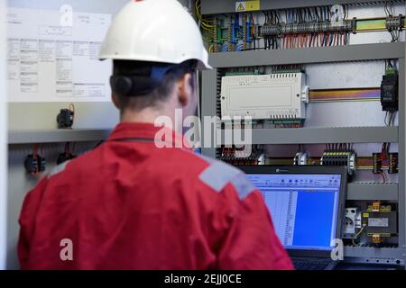 Tema del settore energetico: Un tecnico vestito con tute rosse e un casco bianco che controlla i parametri di riscaldamento su un computer portatile. Apparecchiature di riscaldamento Foto Stock