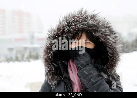 Tempo freddo, donna in maschera medica e cappuccio in pelliccia in piedi sulla strada invernale durante la neve. Concetto di gelo, malattia, febbre, protezione del coronavirus Foto Stock