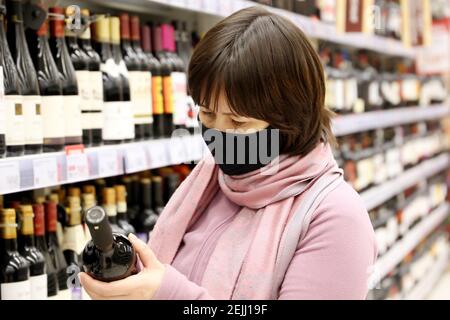 Donna in maschera medica che legge l'etichetta sulla bottiglia di vino rosso. Cliente in negozio di liquori, concetto di scelta e acquisto di alcol Foto Stock