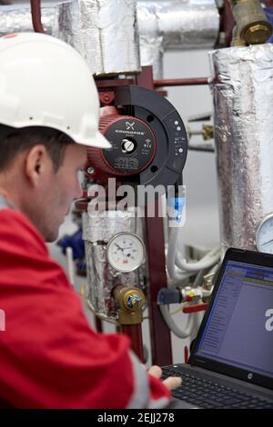 Tema del settore energetico: Un tecnico in rosso e un casco bianco controlla i parametri di riscaldamento su un computer portatile. Manutenzione e ispezione. Foto Stock