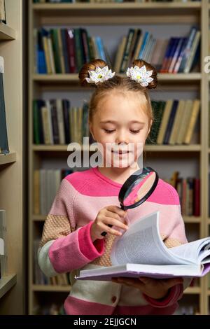 ragazza in abbigliamento casual in biblioteca dopo la scuola, leggendo un'enciclopedia, diligente bambino utilizzare lente di ingrandimento Foto Stock