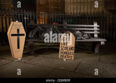 Glasgow, Scozia, Regno Unito. 22 Feb 2021. Nella foto: Una scultura di un senzatetto che dorme su una panchina del parco con un cartello a forma di lapide che recita: "QUI GIACE SCOTLANDS HOMELESS DEATHS". Oggi 23 febbraio 21, il governo scozzese ha rilasciato i dati sui decessi per senzatetto 3 giorni prima, che sono aumentati del 11% rispetto allo scorso anno. Il link https://www.nrscotland.gov.uk/news/2021/homeless-deaths-2019 mostra l'articolo oggi. Credit: Colin Fisher/Alamy Live News Foto Stock