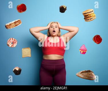 La ragazza grassa nella suite di idoneità vuole iniziare una dieta ma ha dubbi circa il cibo da comprare. Sfondo ciano Foto Stock