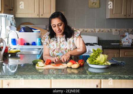 Donna adulta tagliando verdure fresche in cucina - ispanica donna che prepara insalate sane con verdure fresche e biologiche Foto Stock