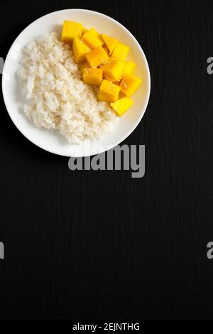 Riso Sticky fatto in casa con semi di mango e sesame su un piatto bianco su una superficie nera, vista dall'alto. Posa piatta, sovratesta, dall'alto. Spazio di copia. Foto Stock