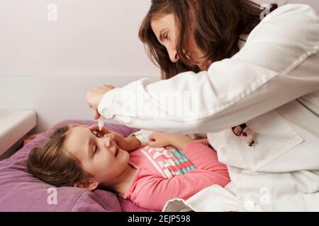 Femmina medico sorridente e dando gocce di occhio ad una bambina nel suo letto. Concetto medico domestico Foto Stock