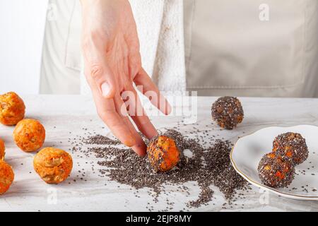 Una chef donna sta preparando i morsi della torta di carote in cucina. Si immerge le palle di carota in un mucchio di semi di chia e rotolarli per rivestire la loro surf Foto Stock