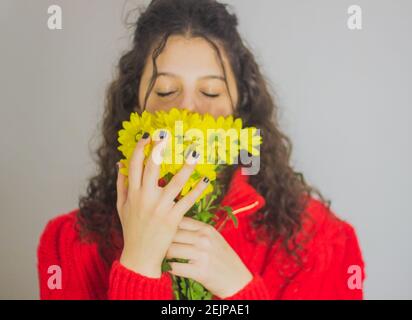 donna brunetta con capelli ricci in maglione rosso che odora il giallo fiori Foto Stock