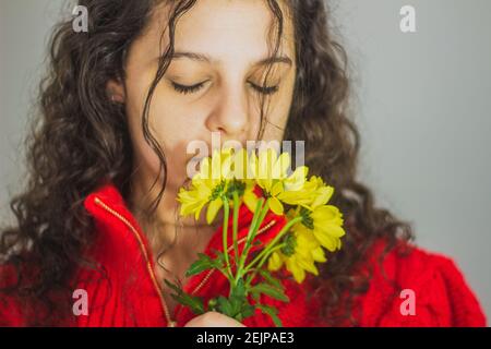 donna brunetta con capelli ricci in maglione rosso che odora il giallo fiori Foto Stock