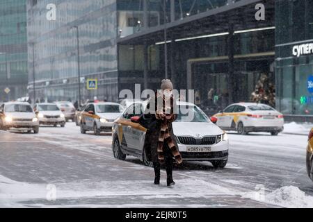 Mosca. Russia. 12 febbraio 2021. Una donna in un cappotto di pelliccia e una maschera medica con uno smartphone in mano cammina in inverno lungo una strada della città contro Foto Stock