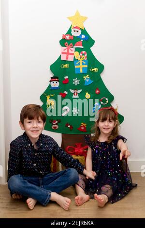 Una bambina carina, dai capelli marroni, dagli occhi blu e un ragazzo carino, dai capelli rossi, dagli occhi blu, vestito in modo intelligente seduto sul pavimento di fronte a un albero di Natale Foto Stock