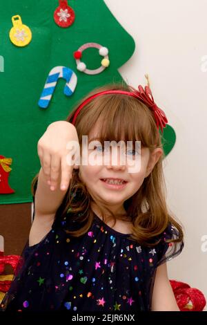 Un bambino carino, dai capelli marroni, dagli occhi blu, vestito con intelligenza sorridente davanti ad un albero di Natale con la sua mano in su Foto Stock